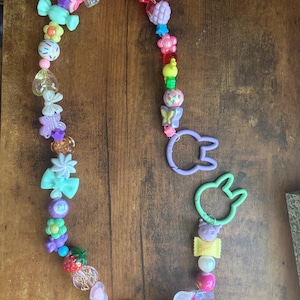 May include: A long beaded lanyard featuring a variety of colorful plastic charms including butterflies, flowers, candy shapes, stars, and hearts. The strand is finished with two light purple and light green rabbit-shaped plastic lobster clasps.