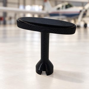 May include: A black T-shaped handle with a textured, wood-grain finish. The handle features a horizontal, oval-shaped grip attached to a vertical cylindrical stem that widens at the base, which includes a small, notched cutout opening.