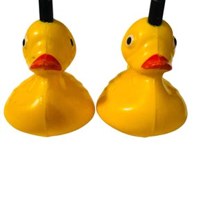 May include: Two yellow rubber ducks with orange beaks and black eyes. Each duck features a small, upright black cylindrical protrusion on the top of its head. The ducks have a smooth, rounded body shape and are positioned side by side.