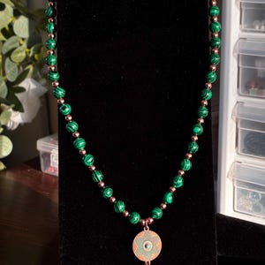 May include: A beaded necklace featuring alternating green malachite spheres and small metallic copper-toned beads. The necklace includes a circular copper-toned pendant with a textured geometric pattern, accented by a small turquoise bead and a copper cone.