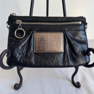 May include: A black leather clutch bag featuring a silver-toned zip closure with a circular pull tab. A rectangular metallic plaque on the front is engraved with the text Coach, Poppy, and Coach est 1941, accented with small heart symbols.