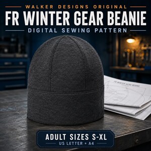 May include: A dark charcoal grey beanie with a multi-panel construction and a wide folded brim. Beside the beanie are printed sewing pattern documents labeled FR WINTER GEAR BEANIE. The text indicates adult sizes S-XL for US Letter and A4.