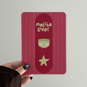 May include: A rectangular card with a dark red vertical striped background. A central dark red oval contains the text "matcha lover" in light green, a light green bowl illustration with small white bunnies and a pink flower, and a light green star.