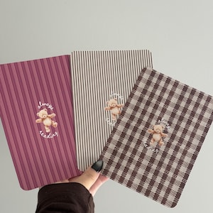 Op de afbeelding: Drie rechthoekige notitieboekjes met een kleine illustratie van een teddybeer in het midden met de tekst "always reading" in cursief schrift. Het eerste notitieboekje heeft verticale magentakleurige strepen, het tweede heeft dunne beige strepen en het derde heeft een bruin ruitpatroon.