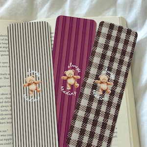 May include: Three rectangular bookmarks featuring a central illustration of a light brown teddy bear with the white cursive text always reading. The left bookmark has thin black stripes, the center has magenta stripes, and the right has a brown plaid pattern.