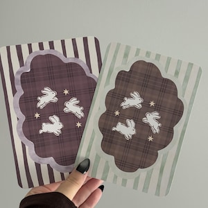 May include: Two rectangular cards featuring a central scalloped shape with a plaid pattern, three white rabbit illustrations, and small stars. One card has a dark purple striped background, while the other has a light green striped background.