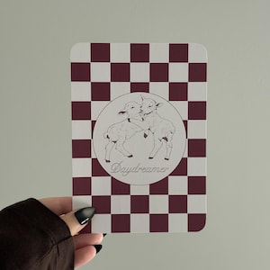 May include: A rectangular card featuring a maroon and white checkerboard pattern. In the centre, a circular white emblem displays a line-art illustration of two lambs embracing, with the word Daydreamer written in cursive script below them.