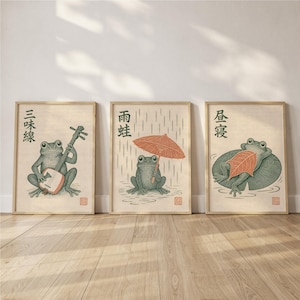 May include: A set of three framed prints featuring green frogs in various activities. The first shows a frog playing a shamisen, the second a frog holding an orange umbrella in the rain, and the third a frog resting on a lily pad with a leaf.