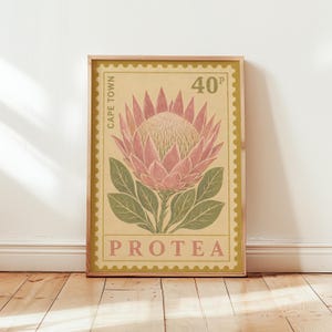 Op de afbeelding: Een rechthoekige wandkunstprint ontworpen als een postzegel met een roze proteabloem met groene bladeren op een lichtgele achtergrond. De tekst luidt CAPE TOWN, 40P en PROTEA in een schreeflettertype. De print heeft een dunne houten lijst.
