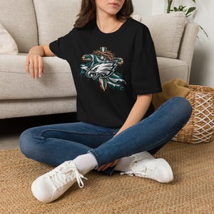 May include: A black short-sleeved t-shirt featuring a graphic print of a stylised eagle head logo layered over a wooden cross wrapped in a crown of thorns, with blue and white fabric accents draped behind the cross design.