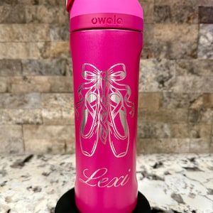 Pode incluir: Uma garrafa de água Owala rosa brilhante com um design gravado em prata de sapatilhas de ballet atadas com um laço. O nome Lexi está gravado em letra cursiva abaixo das sapatilhas. A garrafa tem uma tampa rosa a condizer com fecho.