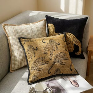 May include: Three square throw pillows. One is beige with a brown grid pattern. Another is black with a large gold leopard silhouette. The third is gold with a repeating black leopard pattern and black piping along the edges.