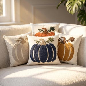 May include: Four square throw pillows with off-white fabric covers. Each pillow features a central punch needle design of a pumpkin in a different color: beige, dark blue, mustard yellow, and orange, all with green stems and curly vines.
