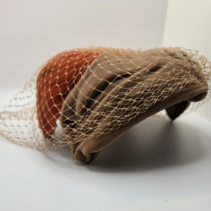 May include: A structured hat featuring a two-tone design with a rust-colored velvet section and a taupe-colored fabric section. The entire piece is covered by a fine, light-colored netting veil that drapes over the front and sides.