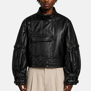 May include: A black leather jacket featuring a high collar, an asymmetrical front button closure, and a large chest pocket. The sleeves are detailed with multiple adjustable buckle straps and small pockets, creating a structured, utility-inspired look.