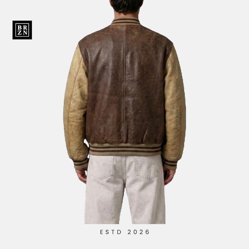 May include: A varsity jacket featuring a dark brown leather body and light tan leather sleeves. The jacket has a ribbed collar, cuffs, and hem with brown and tan stripes. The text BR ZN is in the top left corner and ESTD 2026 is at the bottom.