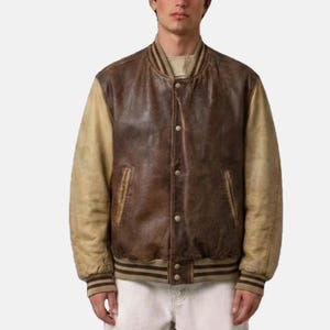 May include: A varsity jacket featuring a dark brown leather body and contrasting tan leather sleeves. The jacket includes a ribbed collar, cuffs, and hem with brown stripes, a snap-button front closure, and two side pockets with leather trim.