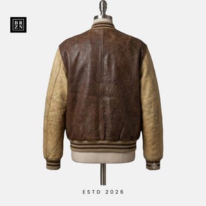 May include: A varsity jacket featuring a dark brown leather body and light tan leather sleeves. The jacket includes a ribbed collar, cuffs, and hem with brown stripes. The text BR ZN appears in a black square in the corner, and ESTD 2026 is below.