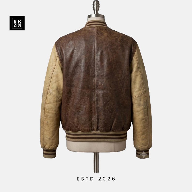 May include: A varsity jacket featuring a dark brown leather body and light tan leather sleeves. The jacket includes a ribbed collar, cuffs, and hem with brown stripes. The text BR ZN appears in a black square in the corner, and ESTD 2026 is below.