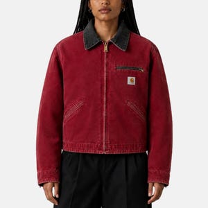 May include: A red canvas jacket featuring a dark gray collar, a full-length front zipper, and long sleeves. The garment includes a horizontal zippered chest pocket on the left side with a small white rectangular Carhartt brand logo patch.