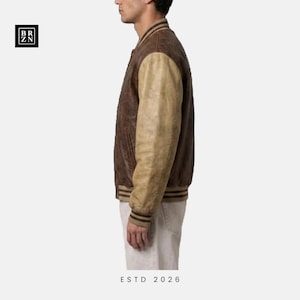May include: A varsity-style jacket featuring a dark brown leather body and contrasting light tan leather sleeves. The garment includes a ribbed collar, cuffs, and hem with brown stripes. A black square logo with the text BR ZN is visible.