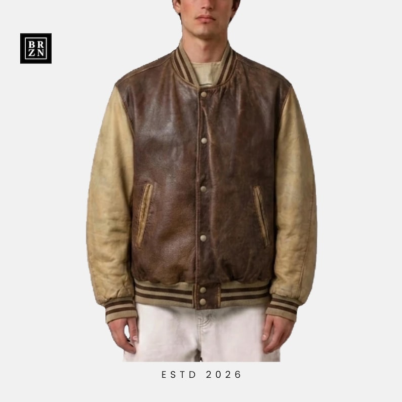 May include: A varsity jacket featuring a dark brown leather body and tan leather sleeves. The jacket has a ribbed collar, cuffs, and hem with brown stripes. It includes a front snap button closure, two side pockets, and the text BR ZN and ESTD 2026.