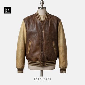 May include: A varsity-style jacket featuring a dark brown leather body and contrasting light tan leather sleeves. It includes a ribbed collar, cuffs, and hem with brown stripes, a snap-button front closure, and two side pockets. The text BR ZN and ESTD 2026 appear.