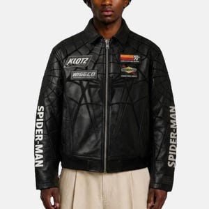 May include: A black leather jacket featuring a debossed spiderweb pattern across the front and sleeves. The jacket has a front zipper, a collar, and various patches including KLOTZ and WISECO. The text SPIDER-MAN is printed on both sleeves.