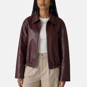May include: A deep burgundy jacket with a smooth finish, featuring a classic collar, a full-length silver zipper, and an elasticized hem. The jacket is worn over a plain white crew-neck t-shirt and light beige trousers with a visible button closure.