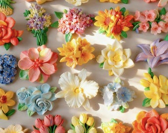 Puede incluir: Una colección de diversos adornos florales tridimensionales con una amplia gama de colores, incluyendo rosa, amarillo, naranja, azul y morado. Cada pieza representa diferentes tipos de flores como rosas, tulipanes y lirios.