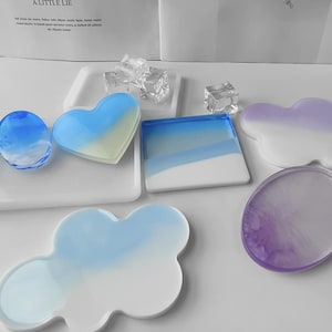 May include: A collection of resin coasters in various shapes including heart, square, cloud, and oval. The coasters feature gradient color transitions in shades of blue, white, and purple. Several clear, cube-shaped resin pieces are scattered among them.