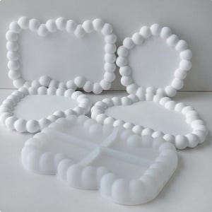 Può includere: Una collezione di stampi in silicone bianco caratterizzati da un bordo in rilievo con texture a bolle. Il set include forme rettangolari e circolari, insieme a uno stampo rettangolare diviso in quattro sezioni, con un bordo sferico uniforme.