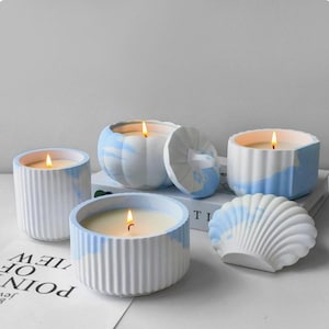 Pode incluir: Quatro velas acesas em recipientes marmorizados brancos e azul-claros. Uma vela está num recipiente cilíndrico canelado, uma num recipiente em forma de abóbora com tampa, uma numa taça canelada e uma num recipiente em forma de concha.