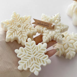Può includere: Tre candele in cera color bianco sporco a forma di fiocco di neve con intricati motivi geometrici. Una candela presenta un cartellino in carta marrone con la scritta HANDMADE stampata in nero. Le candele hanno una finitura liscia e opaca.