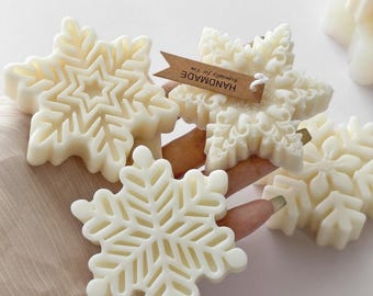 Puede incluir: Tres velas de cera de color blanco roto con forma de copo de nieve y complejos patrones geométricos. Una vela lleva una etiqueta de papel marrón con el texto HANDMADE impreso en negro. Las velas tienen un acabado liso y mate.