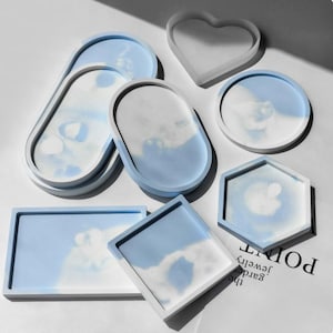 May include: A collection of decorative trays and coasters in various shapes including oval, rectangular, square, hexagonal, and heart-shaped. Each piece features a smooth white border and a marbled light blue and white interior surface.