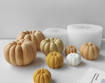 Puede incluir: Una colección de objetos con forma de calabaza en varios tamaños y tonos de naranja, amarillo y blanco, con acabados lisos y texturizados, junto a dos moldes de silicona cilíndricos translúcidos y un libro con el texto Karl Blossfeldt.