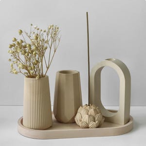 Puede incluir: Un conjunto de hormigón beige que incluye un jarrón cilíndrico acanalado con flores secas, un jarrón facetado, un porta-incienso en forma de loto con varilla y una escultura en arco sobre una bandeja ovalada, todo con acabado mate.