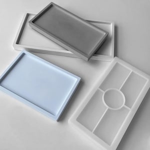 Op de afbeelding: Een collectie rechthoekige dienbladen, waaronder een effen grijs dienblad, een effen wit dienblad, een effen lichtblauw dienblad en een doorschijnende witte siliconen mal met een centraal rond compartiment en vier omliggende rechthoekige secties.