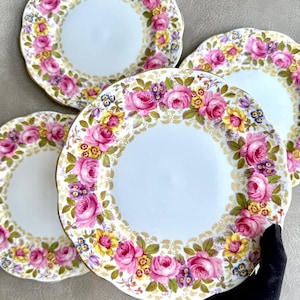 May include: A set of four white ceramic plates with scalloped edges, each featuring a decorative border of pink roses, yellow flowers, and small purple blossoms, accented with gold leaf detailing around the rim and floral pattern.