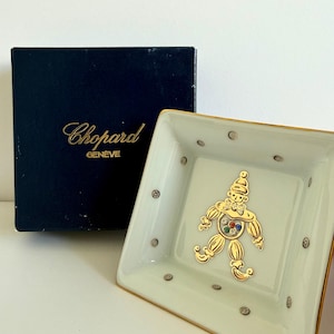 May include: A square white porcelain trinket dish with a gold rim and small grey dots, featuring a central gold clown figure with colourful inset stones. Beside it sits a dark blue box with the gold text Chopard GENEVE printed on the front.