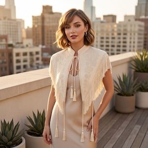 May include: A cream-coloured bolero jacket featuring intricate floral embroidery, a high collar, and long fringe trim along the edges. The garment includes a front tie closure with four hanging tassels, worn over a matching sleeveless satin dress.