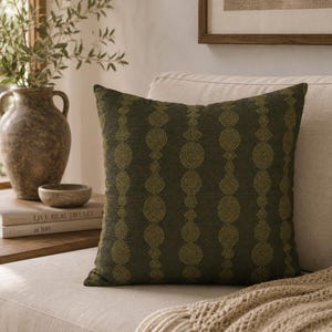 May include: A square throw pillow in a deep olive green color featuring a vertical pattern of repeating textured circles and diamond shapes in a lighter lime green hue. The fabric has a woven appearance with a consistent geometric design.
