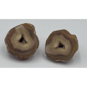May include: Two polished agate slices featuring concentric bands of tan, beige, and brown earth tones. Each stone displays a small, hollow, crystalline center, showcasing natural mineral formations with smooth, rounded edges and a glossy finish.