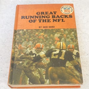 May include: A hardcover book titled Great Running Backs of the NFL by Jack Hand with an orange cover. The cover features a photograph of American football players in action on a field, with an NFL Punt Pass and Kick Library logo in the corner.