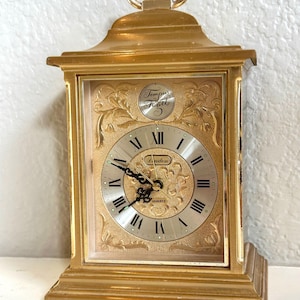 May include: A gold-toned carriage clock with a rectangular frame and a top handle. The clock face features black Roman numerals, ornate gold filigree detailing, and the text Tempus Fugit and Linden. It has black ornate hands and a quartz movement.