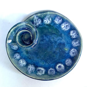 May include: A round ceramic dish with a glossy finish in shades of deep blue and teal. The surface features a raised spiral element on one side and a series of textured, white-flecked blue circular patterns arranged along the outer edge.