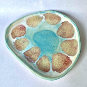May include: A hand-formed ceramic plate with an irregular, rounded triangular shape. The surface features a central pool of bright turquoise glaze surrounded by eight distinct, leaf-shaped impressions in shades of tan, beige, and reddish-brown.