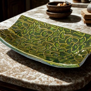 May include: A rectangular ceramic tray with a curved, concave shape. The surface features a vibrant, glossy green glaze with a textured, repeating pattern of overlapping, fan-shaped motifs in a darker shade of green and brown.