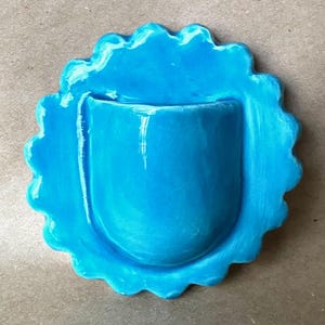 May include: A bright turquoise ceramic wall pocket featuring a scalloped, flower-like edge and a smooth, glossy finish. The central U-shaped pocket is integrated into the circular base, creating a cohesive, solid-colored decorative piece.